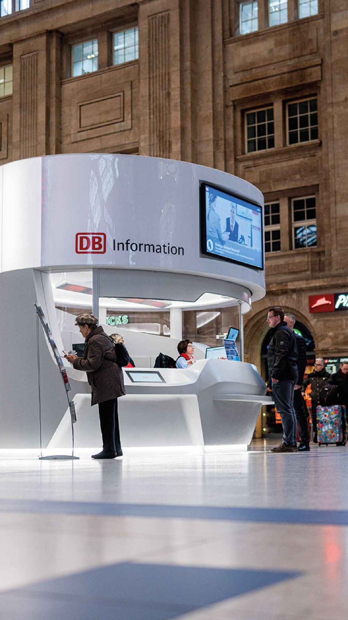 Hochformat-Ansicht-der-runden-DB-Infotheke-aus-Mineralwerkstoff-mit-Mitarbeiter-und-Kunde