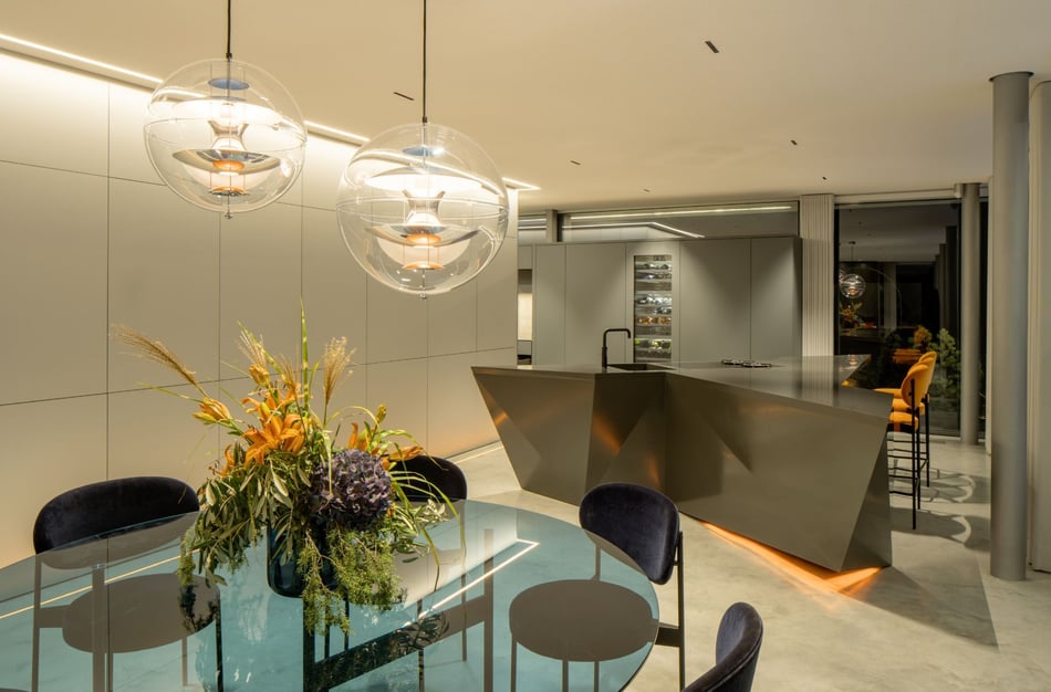 Polygonale-Kuecheninsel-aus-dunklem-Mineralwerkstoff-in-moderner-Wohnumgebung-mit-Designleuchten-und-Esstisch