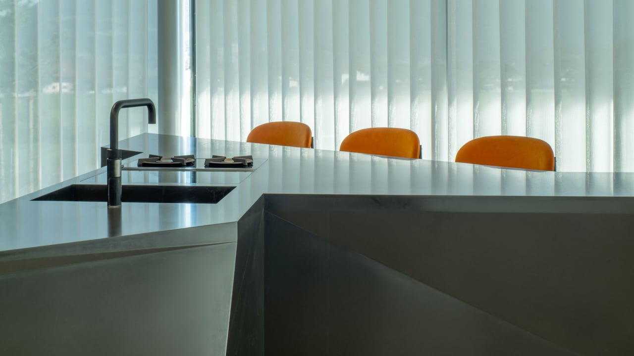 Nahaufnahme-einer-dunklen-Arbeitsplatte-mit-integriertem-Kochfeld-und-Spuele-drei-orange-Stuehle-im-Hintergrund