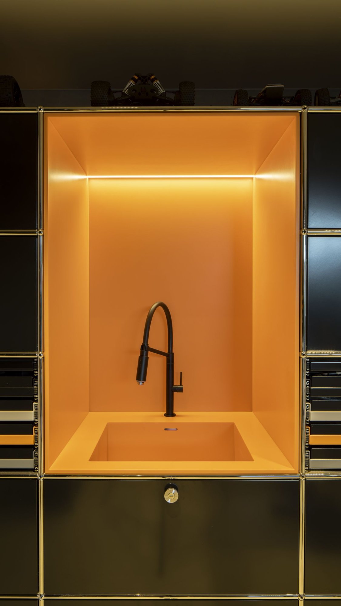 Vertikale-Ansicht-einer-hell-orangefarbenen-Nische-mit-schwarzem-Wasserhahn-und-Spuelbecken-umrahmt-von-einem-schwarzen-Schranksystem Vertikale-Ansicht-einer-hell-orangefarbenen-Nische-mit-schwarzem-Wasserhahn-und-Spuelbecken-umrahmt-von-einem-schwarzen-Schranksystem
