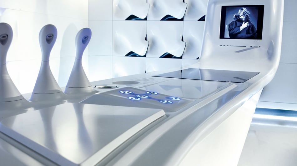 Detailansicht-der-fliessend-geformten-Corian-Kuecheninsel-mit-integriertem-Kochfeld-Bedienelementen-und-drei-skulpturalen-Armaturen-Im-Hintergrund-sind-gewellte-thermisch-verformte-Wandpaneele-zu-sehen