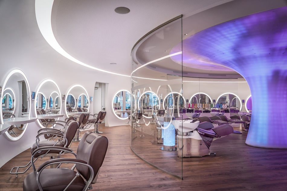 Beleuchteter-Beautysalon-Egger-mit-organisch-geformtem-Mineralwerkstoff-Element-runden-Spiegeln-und-Stuehlen
